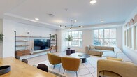Located at 601 N Broad St in Philadelphia's vibrant Northern Liberties neighborhood, this thoughtfully designed single-floor communal space offers a bright and contemporary environment. The open-plan living area is anchored by large windows that flood the room with natural light while framing street-level views of the surrounding cityscape. Light-toned hardwood flooring runs throughout, complementing the clean white walls and creating a warm, welcoming ambiance.

The living area is centered around a plush modular sectional sofa paired with accent chairs in rich mustard tones, arranged around a sleek oval coffee table atop a graphic area rug. A floor-to-ceiling open shelving unit with warm walnut wood and black metal framing houses a large flat-screen television, adding a functional yet stylish focal point. An eye-catching multi-arm brass chandelier serves as a distinctive overhead accent.

A decorative wall featuring arched wood detailing above frosted-glass panels adds architectural character to one side of the room. Adjacent to the living area, a compact kitchenette is outfitted with bold blue cabinetry, open wood shelving, a sink, and small appliances, complemented by a natural wood dining table and seating for group gatherings. The overall aesthetic is cohesive, modern, and inviting.