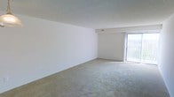 This single-floor space presents a clean, open living area designed with simplicity and functionality in mind. The room features wall-to-wall neutral-toned carpet that extends throughout, providing a soft underfoot feel and a cohesive, understated aesthetic. The walls are painted in a crisp white, creating a bright and airy atmosphere that makes the space feel open and versatile.

The textured ceiling adds a subtle architectural detail, while a ceiling-mounted HVAC vent ensures comfortable climate control year-round. A standout feature of the main living area is the full-height sliding glass door fitted with vertical blinds, which serves as both a source of abundant natural light and a direct connection to the outdoors. The light filtering through this door brightens the interior considerably, enhancing the overall sense of openness.

The layout is straightforward and unobstructed, offering flexibility for a variety of furniture arrangements. The neutral finishes throughout — from the carpet to the walls and ceiling — provide a blank canvas that can accommodate a wide range of interior design styles, making this a highly adaptable living space.