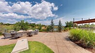 The Grilling Area & Fire Pit at 355 Eldorado Blvd in Broomfield, Colorado, is a thoughtfully designed outdoor amenity space that offers a seamless blend of functionality and natural beauty. The grounds feature expansive stamped concrete patios that provide ample room for outdoor dining and socializing, furnished with multiple seating arrangements including tables and chairs suited for gatherings of various sizes. A handsome wood pergola adorned with string lights anchors a central dining zone, creating a warm and inviting atmosphere for both daytime and evening use. Adjacent to the main patio, a dedicated fire pit area is outfitted with comfortable lounge seating and framed by mature evergreen trees and ornamental grasses, offering a more intimate setting for relaxation. The space is bordered by a wrought iron fence that opens to picturesque views of a nearby pond and the surrounding Colorado landscape. A covered lower-level patio tucked beneath an upper-level deck features exposed wood beam ceilings and wall-mounted lighting, providing a shaded retreat with a cornhole lawn game area on the adjacent green space. The building's contemporary architecture—combining stone masonry, wood siding, and large windows—serves as a striking backdrop, while a steel staircase connects the outdoor levels. The overall ambiance is open, airy, and well-suited for outdoor community living in Colorado's scenic Front Range setting.