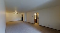 This single-floor space is designed with an open layout that maximizes the sense of roominess throughout the main living area. The living room is finished with wall-to-wall carpet in a neutral tone, providing a comfortable and cohesive foundation underfoot. A ceiling fan is mounted overhead, offering both light and air circulation to the space.

The open floor plan allows for a clear sightline from the living area toward the kitchen, which is partially visible in the background and features dark appliances, including what appears to be a range, along with upper cabinetry. The kitchen is separated from the main living space by a partial wall, creating a subtle division while maintaining the open feel of the layout.

An interior doorway leads to a combined bathroom and laundry area, which is visible from the main living space. This functional zone features wood-toned cabinetry, a vanity, and laundry appliances, making everyday tasks conveniently accessible. The walls throughout are finished in a clean white, contributing to a bright and neutral ambiance. Overall, the space offers a practical and straightforward layout suited for comfortable everyday living.
