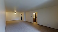 This single-floor space is designed with an open layout that maximizes the sense of roominess throughout the main living area. The living room is finished with wall-to-wall carpet in a neutral tone, providing a comfortable and cohesive foundation underfoot. A ceiling fan is mounted overhead, offering both light and air circulation to the space.

The open floor plan allows for a clear sightline from the living area toward the kitchen, which is partially visible in the background and features dark appliances, including what appears to be a range, along with upper cabinetry. The kitchen is separated from the main living space by a partial wall, creating a subtle division while maintaining the open feel of the layout.

An interior doorway leads to a combined bathroom and laundry area, which is visible from the main living space. This functional zone features wood-toned cabinetry, a vanity, and laundry appliances, making everyday tasks conveniently accessible. The walls throughout are finished in a clean white, contributing to a bright and neutral ambiance. Overall, the space offers a practical and straightforward layout suited for comfortable everyday living.