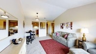 This single-floor residence showcases a thoughtfully designed open-concept layout that seamlessly connects the living area to the kitchen. The living room is highlighted by vaulted ceilings that lend a sense of spaciousness and airiness to the space. Neutral-toned carpeting runs throughout, complemented by warm beige walls that create a cozy and welcoming ambiance. A comfortable gray sofa anchors the living area, accented by decorative throw pillows and a rich burgundy area rug that adds a pop of color and texture to the room. Wall art featuring circular designs in warm tones adds a contemporary decorative touch. A small bistro-style dining table with bar stools provides a casual dining option near the kitchen pass-through. The kitchen, visible through the open pass-through, features warm wood cabinetry and is efficiently laid out for everyday use. A pendant light fixture with a classic bronze finish hangs centrally, providing functional and ambient lighting. Louvered doors offer access to additional spaces, while the overall flow of the floor plan ensures a practical and connected living experience throughout this well-appointed single-level home.