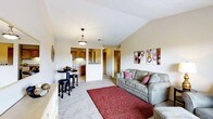 This single-floor residence showcases a thoughtfully designed open-concept layout that seamlessly connects the living area to the kitchen. The living room is highlighted by vaulted ceilings that lend a sense of spaciousness and airiness to the space. Neutral-toned carpeting runs throughout, complemented by warm beige walls that create a cozy and welcoming ambiance. A comfortable gray sofa anchors the living area, accented by decorative throw pillows and a rich burgundy area rug that adds a pop of color and texture to the room. Wall art featuring circular designs in warm tones adds a contemporary decorative touch. A small bistro-style dining table with bar stools provides a casual dining option near the kitchen pass-through. The kitchen, visible through the open pass-through, features warm wood cabinetry and is efficiently laid out for everyday use. A pendant light fixture with a classic bronze finish hangs centrally, providing functional and ambient lighting. Louvered doors offer access to additional spaces, while the overall flow of the floor plan ensures a practical and connected living experience throughout this well-appointed single-level home.