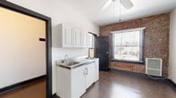 This compact single-floor studio offers a thoughtful blend of industrial character and functional layout. The main living area immediately draws attention with a striking exposed brick accent wall that adds texture and warmth to the otherwise clean, white interior. Dark hardwood floors run throughout the space, grounding the room with a classic, timeless quality. A ceiling fan centered overhead provides both airflow and a casual lighting source, while a large window with blinds invites an abundance of natural light and street-level views, enhancing the sense of openness.

The kitchenette is efficiently arranged along one wall, featuring white upper cabinetry, a stainless steel sink, a compact cooktop, and white base cabinets with under-counter appliance space — providing all the essentials within a streamlined footprint. An electrical panel is conveniently accessible within the main living space. A closet off the main area offers dedicated storage.

The bathroom is clean and functional, appointed with a walk-in tiled shower, a warm wood-toned vanity with a white basin sink, a mirrored medicine cabinet, and a standard toilet — presenting a simple, well-maintained design. Overall, this studio makes efficient use of its footprint while delivering notable architectural character.