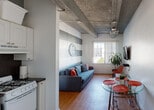 Located at 953 Collins Ave in the heart of Miami Beach, this single-floor unit offers a functional and characterful living space with a distinctive industrial-inspired aesthetic. Throughout the unit, exposed concrete beams define the ceiling, adding architectural depth and a raw, urban character that contrasts with the clean white walls and warm wood flooring found across the main living areas and bedroom.

The living area is bright and welcoming, featuring a window with vertical blinds that allows natural light to fill the room and offers views of the surrounding neighborhood. A wall-mounted flat-screen TV, a blue sofa, and a glass-top dining table with red stools furnish the space, blending practicality with a lively color palette. A bold horizontal stripe accent wall adds visual interest to the room.

The kitchen is efficiently arranged with white cabinetry, dark granite-style countertops, a stainless steel sink, a gas range, and a microwave — providing all the essentials for everyday cooking.

The bedroom is a calm retreat, featuring the same exposed beam ceiling, wood flooring, a window unit for climate control, a closet, and tasteful wall art that reflects the coastal Miami Beach environment.

The bathroom is clean and straightforward, with a combined tub and shower enclosed by a white curtain. This unit presents a practical and characterful option in one of Miami Beach's most vibrant and walkable corridors.
