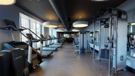 This contemporary fitness center is thoughtfully designed to accommodate a full range of workout disciplines across a single, expansive floor. The main training area is anchored by a row of treadmills and cardio machines positioned directly in front of floor-to-ceiling windows, offering sweeping views of the surrounding urban skyline and filling the space with abundant natural light. Overhead, bold circular drum pendant lights hang from a dramatic dark ceiling, creating a striking contrast that defines the gym's modern aesthetic. Exposed ductwork adds an industrial touch, complementing the sleek, rubber-floored surfaces throughout. The strength training section is generously outfitted with a comprehensive selection of cable machines, selectorized equipment, free weights, and benches, all arranged to maximize usability and flow. A dedicated cycling studio features a row of stationary bikes facing full-length mirrors, warm wood-panel accent walls, and a ballet barre, making it ideal for spin classes or solo cycling sessions. Exercise mats, medicine balls, and storage lockers round out the functional amenities. Mirror installations throughout the space amplify the sense of openness. Together, the facility's design balances high-performance functionality with a polished, upscale ambiance.