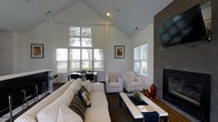 This single-story clubhouse offers an inviting and well-appointed communal space designed for comfort and socializing. The main living area is immediately distinguished by its dramatic vaulted ceiling, which rises to an impressive height and creates a bright, airy atmosphere throughout the room. Large multi-pane windows line the walls, flooding the interior with natural light and offering views of the surrounding greenery outside.

The lounge area is anchored by a sleek, floor-to-ceiling stone-clad fireplace fitted with a wall-mounted flat-screen television above, serving as a striking focal point. Comfortable neutral-toned seating, including a generous sofa and coordinating armchairs, is arranged around a stylish rectangular coffee table, creating an ideal setting for relaxing or socializing.

Adjacent to the main seating area, a dining nook with dark table and chairs provides space for casual meals or meetings. The interior design balances contemporary and transitional styles, featuring clean lines, tasteful artwork, and carefully selected décor accents. Dark hardwood flooring grounds the space and complements the crisp white walls and ceiling throughout.