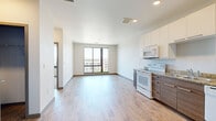 This thoughtfully designed one-bedroom, one-bathroom unit offers a modern living experience across a single floor. The open-concept layout seamlessly connects the living room and kitchen, creating a bright and airy atmosphere enhanced by light wood-style flooring throughout. A striking floor-to-ceiling window in the living area floods the space with natural light while framing panoramic views of the surrounding cityscape and skyline.

The kitchen is efficiently appointed with two-tone cabinetry — white upper cabinets paired with dark wood-grain lower cabinets — complemented by granite-style countertops. Full-size appliances include a range, microwave, dishwasher, and refrigerator, providing everything needed for everyday cooking.

The bedroom is a comfortable, well-proportioned retreat with carpet flooring and neutral walls, offering a quiet separation from the main living area. A dedicated closet provides convenient storage.

The bathroom features a clean, contemporary design with a full soaking tub and shower combination, a dark wood vanity with a granite countertop, and wood-style flooring that ties seamlessly into the rest of the unit. The entryway offers a welcoming transition into the home, with additional closet access nearby. Overall, the unit presents a clean, modern aesthetic ideal for comfortable urban living.
