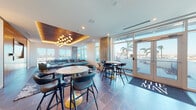 This sophisticated single-floor lounge space, branded "The Man," is designed with a contemporary aesthetic that seamlessly blends comfort and style. The main lounge area is anchored by a striking coffered ceiling with warm amber accent lighting and an eye-catching multi-globe gold chandelier, creating an intimate yet upscale ambiance. Large floor-to-ceiling windows and glass doors flood the space with abundant natural light while offering sweeping views of the surrounding cityscape and palm trees beyond.

The lounge is furnished with a generous L-shaped sectional sofa in soft grey velvet, complemented by a plush green accent armchair, all set atop richly patterned area rugs over large-format slate tile flooring. A flat-screen TV is elegantly recessed into a warm wood-paneled feature wall, adding a media-centric focal point.

The dining and social areas feature round marble-top tables paired with sleek teal leather chairs and bar-height bistro tables with modern stools, providing versatile seating arrangements. A dedicated wet bar or kitchenette area includes navy cabinetry with brass hardware, a white countertop, decorative herringbone tile backsplash, open shelving, and curated wall art, rounding out the space as a fully equipped social hub. Double glass entry doors with branded floor matting complete the polished entrance experience.