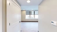Located in the heart of downtown Utica, NY, this thoughtfully designed 2-bedroom, 2-bathroom residence offers a clean, modern aesthetic throughout. The kitchen is a standout feature, showcasing rich dark cabinetry, a white subway tile backsplash, sleek quartz countertops, stainless steel appliances, and a central island with pendant lighting — all complemented by light hardwood-style flooring and a window framing city views. The two bedrooms are comfortably proportioned and carpeted for warmth. The primary bedroom benefits from a sweeping bank of windows offering panoramic downtown views, while the second bedroom features a ceiling fan and its own closet. Three closets throughout the unit provide ample storage. Both bathrooms are well-appointed; one features a tub-shower combination with white subway tile, a bold blue vanity with granite countertop, and large-format tile flooring, while the second includes a walk-in tile shower with matte black fixtures. A connecting hallway ties the layout together efficiently. Soft, neutral tones in sage green and warm gray carry throughout, creating a cohesive and calming ambiance. Recessed lighting and flush-mount fixtures ensure well-lit interiors across every room.