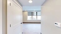 Located in the heart of downtown Utica, NY, this thoughtfully designed 2-bedroom, 2-bathroom residence offers a clean, modern aesthetic throughout. The kitchen is a standout feature, showcasing rich dark cabinetry, a white subway tile backsplash, sleek quartz countertops, stainless steel appliances, and a central island with pendant lighting — all complemented by light hardwood-style flooring and a window framing city views. The two bedrooms are comfortably proportioned and carpeted for warmth. The primary bedroom benefits from a sweeping bank of windows offering panoramic downtown views, while the second bedroom features a ceiling fan and its own closet. Three closets throughout the unit provide ample storage. Both bathrooms are well-appointed; one features a tub-shower combination with white subway tile, a bold blue vanity with granite countertop, and large-format tile flooring, while the second includes a walk-in tile shower with matte black fixtures. A connecting hallway ties the layout together efficiently. Soft, neutral tones in sage green and warm gray carry throughout, creating a cohesive and calming ambiance. Recessed lighting and flush-mount fixtures ensure well-lit interiors across every room.