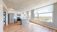 This contemporary single-floor residence combines modern design with functional living. The heart of the home is an open-concept kitchen and living area, where light hardwood flooring extends throughout and large windows flood the space with natural light, framing panoramic views of the surrounding cityscape and treetops beyond. The kitchen is well-appointed with white shaker cabinetry, a white tile backsplash, stainless steel appliances including a refrigerator, electric range, microwave, and dishwasher, and a central island with a clean white countertop. Pendant lighting hangs above the island, complementing the exposed concrete ceiling that lends an industrial character to the space. A stacked washer and dryer unit is conveniently integrated near the kitchen area. The bedroom features plush carpeting, an exposed concrete ceiling, and a large window offering elevated city views. A generously sized walk-in closet with wire shelving provides ample storage. Built-in open shelving near the entry adds both style and practicality. The bathroom is finished with vertically stacked white tile in the tub surround, chrome fixtures, and hardwood flooring. Throughout, the neutral color palette and abundant natural light create a bright, airy, and modern ambiance.