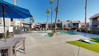 Located in Tempe, Arizona, this outdoor amenity area at 2020 E Willow Creek showcases a resort-style pool and spa complex designed for year-round enjoyment under the abundant Arizona sunshine. The centerpiece is a generously sized swimming pool with crystal-clear turquoise water, accented by decorative blue tile details along the pool floor. Adjacent to the main pool is a separate spa, providing a dedicated relaxation area for residents. The expansive concrete pool deck offers ample space for lounging, furnished with a generous selection of chaise lounge chairs cushioned in blue and complemented by large blue patio umbrellas that provide welcome shade throughout the day. Towering palm trees frame the space, lending a distinct desert resort ambiance. A small patch of manicured turf adds a refreshing touch of greenery. The entire pool area is enclosed by a sleek wrought-iron fence, ensuring both security and an open, airy feel. Surrounding the pool deck, the multi-story residential buildings feature stucco exteriors with tiled rooflines, private balconies, and staircases, all set against the backdrop of a clear blue Arizona sky. The overall atmosphere is vibrant, relaxed, and well-suited for outdoor living.