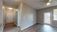 Located at 1803 Marshall Cross in San Antonio, TX, this single-story unit offers a practical and well-organized layout designed for comfortable everyday living. The living area features durable wood-look flooring and vaulted ceilings with a ceiling fan, creating an open and airy atmosphere. A glass-paneled door floods the space with natural light and provides outdoor views. The kitchen is efficiently arranged in an L-shape configuration, equipped with light wood cabinetry, dark countertops, and black appliances including an electric range with overhead ventilation, a dishwasher, and a sink. The wood-look flooring continues throughout this area for easy maintenance. A central hallway connects the living spaces to the bedrooms, each finished with plush carpeting and windows fitted with blinds that allow natural light to filter in. The bedrooms are generously proportioned and offer a calm, neutral atmosphere. The unit includes two bathrooms: one features a vanity with light wood cabinetry and dark countertops paired with wood-look flooring, while the other includes a tub-shower combination with tile surround. Neutral gray walls throughout the unit provide a clean, cohesive aesthetic well-suited to a variety of interior styles.
