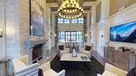 This sophisticated single-floor space makes a striking first impression with its dramatic double-height ceilings and refined architectural detailing. Coffered ceiling panels and crisp white columns frame the room, lending a sense of classical structure and grandeur. At the heart of the space, a large two-tiered candle-style chandelier serves as a commanding focal point, casting warm, ambient light throughout. A floor-to-ceiling stone fireplace anchors one side of the room, adding texture and a cozy, inviting atmosphere. The seating area is thoughtfully arranged with wingback chairs and a stone-topped coffee table set atop a richly patterned area rug, creating an intimate gathering space. Expansive multi-pane windows, flanked by bold drapery panels, draw in generous natural light and offer views to the exterior, enhancing the open, airy feel of the interior. Wall sconces and large-scale artwork — including a dramatic landscape painting — contribute to the curated, upscale aesthetic. The hardwood flooring, neutral color palette, and carefully selected furnishings all work together to create a polished, welcoming environment that balances elegance with comfort. This space is well-suited for relaxed socializing or quiet reflection.