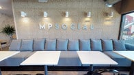 MP Social is a thoughtfully designed social gathering space that seamlessly blends comfort, style, and function across a single open floor. The entrance welcomes visitors with a striking lounge area anchored by a large-format marble-look tile accent wall, plush dark sofas, natural wood stump coffee tables, and sculptural geometric floor lamps—creating an inviting and contemporary atmosphere.

The main gathering area features light wood flooring, exposed industrial ductwork overhead, and an array of café-style seating including bistro tables with black bentwood chairs. A signature banquette runs along the textured feature wall boldly branded with the "MP Social" name, illuminated by wall-mounted accent lights.

The service bar is a standout element, stretching across the back of the space with a vivid blue-toned distressed panel facade, white quartz countertop, and bar stools. The kitchen area behind supports food service with ample cabinetry and equipment, and writable column surfaces add a playful, communal touch.

A bar-top seating nook with large windows overlooks the outdoor terrace, which offers two distinct zones: a dining area with café tables surrounded by lush planters, and a lounge zone furnished with weather-resistant sofas and chairs. The terrace provides open sky views and abundant natural light, making it an ideal setting for relaxed social gatherings.
