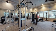 This spacious fitness center is designed to accommodate a wide variety of workout styles and training needs. The interior features a clean, modern aesthetic highlighted by shiplap wall accents, crown molding, and neutral tones that create a polished yet functional environment. Durable carpet tile flooring covers the workout area throughout, providing both traction and comfort underfoot.

The facility is comprehensively equipped for strength training, with a multi-station cable machine, a dedicated free weight area stocked with a full rack of dumbbells, weight benches, and a functional training rig complete with a heavy bag and TRX suspension system. A tire for functional fitness training adds a dynamic element to the floor space.

The cardio section includes multiple treadmills, ellipticals, a stair climber, and a stationary bike, each positioned near large windows that allow natural light to fill the room and offer outdoor views. Mounted flat-screen televisions are positioned throughout for entertainment during workouts.

Ceiling fans and pendant light fixtures hang from the elevated tray ceilings, contributing to the room's open, airy atmosphere. Full-length mirrors along the walls visually expand the space and provide functional feedback for lifters. Double glass entry doors with built-in shelving near the entrance complete the well-organized, welcoming layout.