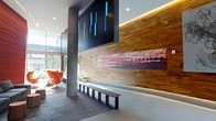 This single-floor lobby space showcases a thoughtfully designed interior that blends contemporary aesthetics with warm, inviting materials. The focal point of the space is a striking wood-planked accent wall finished in rich, warm tones, which serves as a backdrop for a large, vivid piece of abstract artwork rendered in deep reds and oranges, adding a bold visual anchor to the room. Above, a sleek black panel displays illuminated blue vertical light elements, lending a modern, tech-forward identity to the space.

The flooring transitions between carpet and hard surfaces, defining distinct zones within the lobby. A minimalist bench with dark metal legs is positioned along the accent wall, offering functional seating while maintaining the clean design language. In the background, a seating area features iconic orange chair designs that introduce a pop of color and a nod to mid-century modern style.

Exposed concrete columns provide an industrial contrast to the warmth of the wood paneling, while recessed and cove lighting cast a soft, ambient glow throughout. Large windows along the perimeter allow natural light to filter in, enhancing the open, welcoming atmosphere of the space.