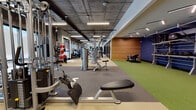 This professionally designed fitness center offers a comprehensive workout environment spread across a single expansive floor. The space is defined by an industrial aesthetic, featuring exposed concrete ceilings with sleek linear lighting that illuminates the facility throughout.

The main floor is thoughtfully divided into distinct training zones. A dedicated cardio area lines one side of the facility, equipped with treadmills, ellipticals, and stair climbers, all positioned alongside large floor-to-ceiling windows that allow ample natural light to fill the space. A glass-partitioned private training studio runs along the opposite side, featuring hardwood flooring and providing a more intimate setting for focused sessions.

The strength training zone is outfitted with a comprehensive selection of cable machines, selectorized equipment, and multi-functional benches. A free weights area includes a full rack of dumbbells and an adjustable bench, positioned near large windows with exterior views.

The functional training zone stands out with vibrant green turf flooring set against bold navy blue accent walls. This area is stocked with wall-mounted storage racks holding medicine balls, kettlebells, slam balls, and battle ropes, alongside a power rack for barbell training. Warm wood-panel wall accents add a refined touch to this dynamic space, creating an energetic yet polished training environment.