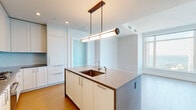 Located in Chicago's prestigious Streeterville neighborhood, this high-rise residence at 514 N Peshtigo Ct offers a refined living experience defined by modern design and breathtaking views. The open-concept layout is anchored by a sleek kitchen featuring floor-to-ceiling white shaker cabinetry, a soft blue subway tile backsplash, quartz countertops, and stainless steel Bosch appliances including a gas cooktop and wall oven. A substantial center island with an undermount sink and a striking tubular brass-accented pendant light serves as both a functional workspace and a visual centerpiece. Light hardwood flooring flows seamlessly throughout the living areas and bedrooms, enhancing the bright, airy atmosphere. The living space is framed by expansive corner windows that frame sweeping panoramic views of Lake Michigan and the Chicago skyline, flooding the interior with natural light. The bathroom is elegantly appointed with herringbone marble tile flooring, subway tile walls, a marble-topped vanity with gold hardware, an illuminated mirror, and a glass-enclosed walk-in shower. A well-proportioned hallway with louvered closet doors with gold hinges provides ample storage. Clean white walls, recessed lighting, and a consistently modern palette complete this polished, luminous residence.