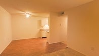This single-floor space offers an open, functional layout that connects the living area directly to the kitchen. The living room features wood-style laminate flooring that transitions to carpet, creating distinct zones within the open plan. A ceiling fan with a built-in light fixture is mounted overhead, providing both air circulation and ambient lighting. The textured ceiling adds a subtle architectural detail throughout the space.

The kitchen is partially open to the living area, separated by a partial wall that defines each space while maintaining an airy feel. White cabinetry lines the kitchen walls, offering ample storage, and is complemented by a countertop with a built-in sink. The white cabinet finish gives the kitchen a clean, classic appearance.

An HVAC vent is positioned high on the wall, ensuring efficient climate control throughout the unit. The neutral wall tones in warm beige create a cohesive, understated palette that allows for versatile interior styling. Overall, the space presents a straightforward, practical layout suited for comfortable everyday living.
