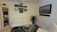 This single-floor space embraces a clean, modern aesthetic throughout its living area. The room is anchored by a white faux-leather futon sofa, offering both style and versatility, paired with a round glass-top coffee table supported by a sculptural chrome base. A plush, dark charcoal area rug grounds the seating arrangement and adds a layer of texture and contrast against the light-toned flooring.

The walls are kept neutral with a crisp off-white finish, allowing the décor to stand out effortlessly. A striking multi-panel black-and-white photographic art piece depicting a dock extending over calm water serves as a bold focal point on one wall, while a flat-screen television is wall-mounted on the adjacent wall for an uncluttered, streamlined look. A potted tropical plant introduces a touch of natural greenery, softening the overall minimalist atmosphere.

Overhead, a pendant light fixture hangs from the ceiling, providing both functional illumination and a subtle design element. The space maintains a cohesive monochromatic color palette of whites, blacks, and grays, creating an atmosphere that feels contemporary, calm, and well-curated. Overall, the layout is efficient and thoughtfully arranged, making excellent use of the available space.