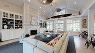 This single-floor clubhouse showcases a sophisticated coastal aesthetic throughout its expansive, open layout. The centerpiece of the space is a grand great room with soaring vaulted ceilings that create an airy, open atmosphere. A striking white-paneled fireplace with a marble surround anchors one wall, flanked by built-in cabinetry and display shelving accented in teal — a nod to the nautical theme. Generous sectional sofas in neutral tones, accented with navy and orange throw pillows, are arranged around glass-topped coffee tables, encouraging relaxed gathering. Natural light pours in through a wall of large, multi-pane windows and French doors, offering views of the lush surroundings. A suspended wooden canoe serves as a dramatic overhead installation, adding an artful, waterfront-inspired focal point. Ceiling fans with pendant lighting complement the vaulted space. The kitchen area features dark cabinetry, quartz countertops, stainless steel appliances, and a beadboard island with bar seating — ideal for entertaining. Light hardwood flooring runs through the kitchen and entry zones, transitioning to plush carpeting in the lounge area. The 3D overview reveals an additional billiards and dining area, rounding out this well-appointed, community-oriented space.