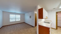 This single-floor unit offers a practical and well-organized layout designed to maximize functionality. The open living area is finished with neutral wall-to-wall carpeting and crisp white walls, creating a clean, versatile backdrop. A window allows natural light to filter into the space, offering views of the surrounding neighborhood. The living area connects seamlessly to the kitchen via a half-wall partition, providing an open sightline between the two spaces.

The galley-style kitchen is efficiently arranged with white cabinetry on both sides, warm wood-trimmed upper and lower cabinets, light countertops, and durable tile-style flooring. White appliances including an electric range and refrigerator are included. A doorway from the kitchen leads to a dedicated pantry or storage room with wire shelving, adding notable storage capacity.

The bedrooms are carpeted and well-lit, each featuring windows with blinds. One bedroom includes a large sliding-door closet, while another offers a generously sized walk-in closet outfitted with wire shelving along multiple walls. The bathroom is thoughtfully designed with accessibility in mind, featuring grab bars near the toilet and walls, a vanity with storage, and light-toned flooring. Overall, the unit presents a clean, functional living environment.