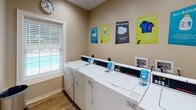 This single-floor amenity space is thoughtfully designed to offer residents two distinct, functional areas that enhance everyday living. The entrance greets visitors with a clean, neutral-toned foyer finished with warm wood-look flooring and crisp white trim, leading into the suite's core amenities.

The standout feature is the "Pup Wash" — a dedicated pet grooming and bathing area branded exclusively for residents. Clad in white subway tile from floor to ceiling, this cheerful space is equipped with a stainless steel wash basin, a step ramp for easy pet access, and drainage built directly into the flooring. Playful paw print decals and informational signage adorn the walls, creating a welcoming and practical environment for pet owners. Protective aprons and waste disposal bins are conveniently provided within the space.

Adjacent to the pet wash area is a well-appointed laundry room featuring multiple full-sized commercial washers and dryers arranged in a neat row. The room is brightened by a window with blinds, allowing in natural light, and is complemented by colorful laundry tip posters that add a lively touch to the space. The consistent use of wood-look flooring throughout ties both areas together, creating a cohesive and polished amenity suite.