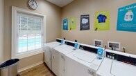This single-floor amenity space is thoughtfully designed to offer residents two distinct, functional areas that enhance everyday living. The entrance greets visitors with a clean, neutral-toned foyer finished with warm wood-look flooring and crisp white trim, leading into the suite's core amenities.

The standout feature is the "Pup Wash" — a dedicated pet grooming and bathing area branded exclusively for residents. Clad in white subway tile from floor to ceiling, this cheerful space is equipped with a stainless steel wash basin, a step ramp for easy pet access, and drainage built directly into the flooring. Playful paw print decals and informational signage adorn the walls, creating a welcoming and practical environment for pet owners. Protective aprons and waste disposal bins are conveniently provided within the space.

Adjacent to the pet wash area is a well-appointed laundry room featuring multiple full-sized commercial washers and dryers arranged in a neat row. The room is brightened by a window with blinds, allowing in natural light, and is complemented by colorful laundry tip posters that add a lively touch to the space. The consistent use of wood-look flooring throughout ties both areas together, creating a cohesive and polished amenity suite.