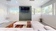 Located at 3480 Todos Santos St in Santa Fe, New Mexico, this single-floor space offers an efficient and thoughtfully designed open-concept layout that seamlessly integrates the living, dining, and kitchen areas. Throughout the space, large-format hexagonal tile flooring in a neutral gray tone establishes a cohesive and modern foundation.

The living area is bright and airy, featuring crisp white walls, recessed lighting, and multiple horizontal windows that invite natural light and provide views of the surrounding neighborhood. A bold, deep-green slatted accent wall serves as a striking focal point, anchoring a flat-screen TV and adding a distinctive architectural element to the room. Comfortable seating is arranged over a vibrant, traditional-style area rug, creating a warm contrast against the contemporary backdrop.

The dining area flows naturally from the living space, accommodating a full dining table and chairs alongside a woven area rug. Additional windows continue to bring in natural light, enhancing the open feel of the combined space.

The kitchen is well-appointed with warm wood-grain cabinetry, open upper shelving, and white quartz countertops. A standout hexagonal tile backsplash in varying shades of gray adds visual interest, while stainless steel appliances — including a range and refrigerator — complete the functional and stylish cooking space.