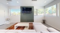 Located at 3480 Todos Santos St in Santa Fe, New Mexico, this single-floor space offers an efficient and thoughtfully designed open-concept layout that seamlessly integrates the living, dining, and kitchen areas. Throughout the space, large-format hexagonal tile flooring in a neutral gray tone establishes a cohesive and modern foundation.

The living area is bright and airy, featuring crisp white walls, recessed lighting, and multiple horizontal windows that invite natural light and provide views of the surrounding neighborhood. A bold, deep-green slatted accent wall serves as a striking focal point, anchoring a flat-screen TV and adding a distinctive architectural element to the room. Comfortable seating is arranged over a vibrant, traditional-style area rug, creating a warm contrast against the contemporary backdrop.

The dining area flows naturally from the living space, accommodating a full dining table and chairs alongside a woven area rug. Additional windows continue to bring in natural light, enhancing the open feel of the combined space.

The kitchen is well-appointed with warm wood-grain cabinetry, open upper shelving, and white quartz countertops. A standout hexagonal tile backsplash in varying shades of gray adds visual interest, while stainless steel appliances — including a range and refrigerator — complete the functional and stylish cooking space.