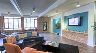 Located in Amarillo, TX, this single-floor space showcases a thoughtfully designed interior that blends rustic and contemporary elements. The main living area is anchored by stacked stone accent walls that add texture and warmth throughout the space. Large windows along one wall invite abundant natural light, offering views of the surrounding greenery while brightening the interior.

The lounge area is furnished with a sleek dark leather sectional sofa accented with colorful throw pillows, complemented by an orange accent chair and a dark coffee table topped with reading materials. A large area rug grounds the seating arrangement, creating a defined and comfortable gathering space.

A standout feature is the bold teal accent wall, which houses a flat-screen television and decorative shelving displaying curated accessories and a potted tree, adding a lively pop of color. Track lighting along the ceiling provides targeted illumination throughout. A secondary seating nook near the windows offers an additional area for relaxation. The large-format tile flooring runs throughout, tying the space together with a clean, durable finish that complements the overall modern-rustic ambiance.