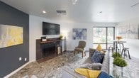 Located at 11239 SE 260th St in Kent, Washington, this thoughtfully designed single-floor space blends contemporary style with functional, communal living. The open layout is anchored by a striking kitchen area featuring dark cabinetry, a large waterfall-edge marble-look island with bar seating, stainless steel appliances, and a white hexagonal tile backsplash accented by rustic open wood shelving. Pendant lighting in matte black adds a modern touch, while light hardwood floors and bold navy-blue accent walls create a vibrant, welcoming atmosphere. A bright yellow accent wall adds a pop of energy to the space.

The lounge area offers a comfortable, residential feel with a dark brick fireplace topped by a wooden mantle, a wall-mounted television, and large windows that frame views of the surrounding greenery. Soft, neutral furnishings with blue and yellow accent pillows complement the overall color palette.

An upper mezzanine level overlooks the main floor, revealing soaring ceilings, exposed ductwork, dark wood structural columns, and floor-to-ceiling windows that flood the interior with natural light. The industrial-modern aesthetic is balanced by warm finishes and carefully curated décor, making this a versatile and inviting gathering space.