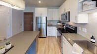This well-designed one-bedroom, one-bathroom residence offers a thoughtfully organized single-floor layout that maximizes functionality and comfort. The open-concept living area features light wood-style flooring, neutral wall tones, and large floor-to-ceiling windows with black-framed French doors that lead to a private balcony, flooding the space with natural light and providing views of the surrounding community.

The kitchen is a standout feature, equipped with a full suite of stainless steel appliances including a refrigerator, range, and over-the-range microwave. Crisp white shaker cabinetry, grey quartz countertops, a subway tile backsplash, and an extended kitchen island create a clean, contemporary aesthetic ideal for both cooking and casual dining.

The bedroom is generously proportioned, featuring plush carpeting and oversized windows that frame outdoor views, enhancing the sense of openness. A roller shade provides flexible light control.

The bathroom offers a clean, modern design with a soaking tub and shower combination, wood-look tile flooring, and a white vanity with ample counter space. Notably, the unit includes a convenient in-unit stacked washer and dryer tucked neatly within a dedicated closet adjacent to the bathroom, adding a practical and desirable amenity to everyday living.