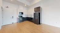 This contemporary single-floor unit is designed with a clean, modern aesthetic and a practical open layout. Warm-toned hardwood-style flooring runs seamlessly throughout the entire space, creating a cohesive and inviting atmosphere. Crisp white walls and recessed lighting contribute to a bright, airy feel.

The open-plan living area flows directly into the kitchen, making the space feel connected and functional. A large window in the living area frames an urban outdoor view, allowing ample natural light to fill the room. The kitchen is outfitted with a striking two-tone cabinetry scheme — dark lower cabinets contrasted with white upper cabinets — paired with light countertops and black appliances, including a refrigerator, electric range, and built-in microwave. A single-basin sink completes the setup.

The bedroom is a calm, well-proportioned retreat with the same consistent flooring and neutral white walls, offering a quiet and versatile space. The bathroom features a full tub-shower combination with white tile surround, a toilet, and a vanity with an integrated sink and mirror, all finished in a clean, minimalist style. The consistent design language across all rooms gives the unit a polished and cohesive character throughout.