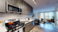 This well-appointed single-floor residence showcases a thoughtfully designed layout with modern finishes throughout. The open-concept kitchen is a standout feature, outfitted with dark wood-grain cabinetry extending to the ceiling, stainless steel appliances including a gas range, built-in microwave, refrigerator, and dishwasher, complemented by light quartz countertops and a sleek subway tile backsplash. Light hardwood flooring runs through the kitchen and living areas, creating a seamless and cohesive flow.

The living area receives generous natural light through large windows dressed with clean white blinds, giving the space an airy, open feel. A dining area sits adjacent, providing a functional and inviting space for meals and entertaining.

The bedroom offers a cozy and curated atmosphere, highlighted by a bold herringbone-patterned accent wall, bright windows that allow ample natural light, and practical storage solutions. A dedicated walk-in closet area with built-in shelving and hanging space connects conveniently to the bedroom.

The full bathroom is finished with large-format neutral tiles, a soaking tub with shower, and dark wood-grain vanity cabinetry, maintaining the modern aesthetic consistent throughout the residence.