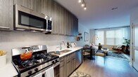 This well-appointed single-floor residence showcases a thoughtfully designed layout with modern finishes throughout. The open-concept kitchen is a standout feature, outfitted with dark wood-grain cabinetry extending to the ceiling, stainless steel appliances including a gas range, built-in microwave, refrigerator, and dishwasher, complemented by light quartz countertops and a sleek subway tile backsplash. Light hardwood flooring runs through the kitchen and living areas, creating a seamless and cohesive flow.

The living area receives generous natural light through large windows dressed with clean white blinds, giving the space an airy, open feel. A dining area sits adjacent, providing a functional and inviting space for meals and entertaining.

The bedroom offers a cozy and curated atmosphere, highlighted by a bold herringbone-patterned accent wall, bright windows that allow ample natural light, and practical storage solutions. A dedicated walk-in closet area with built-in shelving and hanging space connects conveniently to the bedroom.

The full bathroom is finished with large-format neutral tiles, a soaking tub with shower, and dark wood-grain vanity cabinetry, maintaining the modern aesthetic consistent throughout the residence.