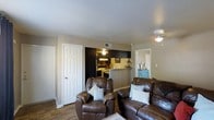 This single-floor, one-bedroom, one-bathroom unit offers a practical and comfortable layout designed for easy living. The living area serves as the central gathering space, featuring wood-style flooring and a ceiling fan overhead. The open sightlines connect the living room directly to the kitchen, creating a sense of flow and openness throughout the common areas.

The kitchen is equipped with dark cabinetry that contrasts with the lighter walls, and includes a gas range, refrigerator, and overhead lighting. A peninsula or pass-through counter separates the kitchen from the living space, providing additional prep or dining surface area while maintaining visual connectivity between rooms.

Multiple doorways lead from the living area to other parts of the unit, offering access to what appear to be a closet and additional rooms. The neutral wall tones throughout the space provide a versatile backdrop suitable for a variety of décor styles. Overall, the unit presents a compact yet functional living environment with a straightforward, single-level layout and utilitarian design elements that prioritize ease of use and comfort.