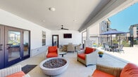 Located at 4406 Helen Rd in Jeffersonville, Indiana, this covered outdoor amenity space serves as a stylish and functional community lounge area positioned directly adjacent to a resort-style swimming pool. The single-level structure features a clean white shiplap exterior with white-painted brick accents along the lower walls, giving the space a bright, modern aesthetic that complements the surrounding residential buildings.

The covered patio is generously proportioned, providing ample room for both lounging and dining. A central stone fire pit bowl serves as a focal point for the seating area, which is furnished with wicker chairs featuring bold orange cushions and accompanying side tables. A full dining set with wicker chairs and a round wooden table offers a dedicated space for outdoor meals. Ceiling fans are installed overhead to ensure comfort during warm Indiana summers, while a wall-mounted flat-screen television adds an entertainment element to the space.

A striking stone fireplace column anchors one end of the patio, adding architectural character and warmth. Large glass double doors open directly toward the pool deck, seamlessly connecting the covered lounge to the sunny outdoor pool area, where lounge chairs, umbrellas, and additional seating are visible. The open-air design allows abundant natural light and unobstructed views of the pool and surrounding community buildings, creating an inviting, resort-inspired atmosphere ideal for year-round outdoor living.