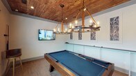 This dedicated game room combines rustic charm with refined design, creating an inviting space ideal for entertainment and recreation. The centerpiece of the room is a full-size pool table with a rich wood frame and blue felt surface, complemented by two striking wagon-wheel style chandeliers suspended from a stunning natural wood-plank ceiling. The warm tones of the cedar-style ceiling create a cozy, lodge-inspired ambiance that contrasts beautifully with the crisp white walls. A wall-mounted flat-screen television ensures the space doubles as a media lounge, while decorative framed artwork adds a polished finishing touch. The lower portion of the walls features classic wainscoting-style paneling, lending an air of architectural sophistication. Light wood-tone flooring ties the room's elements together seamlessly, providing a neutral foundation that enhances the warm, welcoming atmosphere. Recessed lighting supplements the chandeliers, ensuring the room is well-lit for gameplay. This thoughtfully designed game room offers a perfect balance of rustic character and modern amenities, making it an exceptional space for socializing and relaxation.