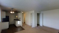 This single-floor unit presents an open-concept layout that integrates the kitchen and living area into one cohesive space. The kitchen is positioned along one wall and features white cabinetry, a dark countertop with a double-basin sink, and a ceiling fan overhead, providing both functionality and airflow. The flooring transitions from dark hardwood in the kitchen area to wall-to-wall carpeting in the main living space, clearly delineating the two zones.

The living area is generously sized and benefits from a neutral color palette of white walls and light carpet, contributing to a clean and airy feel. A textured popcorn ceiling runs throughout the unit, consistent with classic construction detailing. Two closets are visible along the back wall — one a bifold-door closet with built-in shelving and another a standard hinged-door closet with a hanging rod and shelf — offering ample storage options. A hallway visible in the background suggests access to additional rooms, such as a bathroom or bedroom, beyond the main living space.