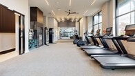 Located at 3068 Briarcliff Rd NE in Atlanta, Georgia, this well-appointed fitness center offers a comprehensive exercise facility across a single floor. The space is thoughtfully divided into distinct workout zones, each designed to support a variety of training styles.

The cardio area features a row of modern treadmills and elliptical machines equipped with digital screens, all positioned alongside large, multi-pane windows that flood the area with natural light and offer views of the surrounding greenery. A ceiling fan overhead aids in ventilation and comfort during workouts.

The strength training zone is a standout feature, boasting soaring double-height ceilings framed by expansive floor-to-ceiling windows that create an open, airy atmosphere. This section is outfitted with a power rack, a full dumbbell rack, weight plates, plyo boxes, and adjustable benches — catering to a wide range of strength and functional training needs.

A dedicated cable machine station and a textured accent wall with dark tile provide visual interest and variety to the space. Full-length mirrors are strategically placed to enhance depth perception and assist with form during exercises.

The facility also includes a water fountain for convenience. Recessed linear lighting and track lighting throughout create a clean, modern aesthetic that is both functional and visually engaging.