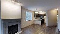 This single-floor space showcases an open-concept design that seamlessly connects the living area and kitchen, creating a cohesive and functional layout. The living area is anchored by a tile-surround fireplace, adding a warm focal point to the room. Hardwood-style flooring runs throughout, lending a clean, contemporary look that ties the spaces together.

A large window allows natural light to fill the living area, complemented by a track lighting fixture overhead. The kitchen is well-appointed with dark espresso-toned cabinetry, stainless steel appliances including a range, microwave, and refrigerator, and a raised breakfast bar that provides additional seating and counter space. A pendant light fixture adds a stylish touch above the kitchen area, while under-cabinet and overhead lighting ensure the workspace is well-lit.

The neutral wall tones and warm flooring create a balanced, inviting ambiance throughout. The open flow between the kitchen and living space makes the unit feel expansive and practical for everyday living.