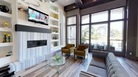 This contemporary lounge space makes an immediate impression with its dramatic ceiling height and thoughtfully curated design. The focal wall showcases a striking combination of a linear fireplace, a wall-mounted flat-screen television, and built-in open shelving adorned with decorative accents — all framed by bold marble-effect paneling that adds texture and visual depth. The flooring features wide-plank wood-look tile in a cool, multi-toned palette that ties the room's neutral and earthy tones together seamlessly. Plush, mustard-gold upholstered accent chairs are arranged around a sleek glass-top coffee table, creating an inviting conversation area. A cushioned sofa in a complementary tone rounds out the seating arrangement, offering comfort without compromising the sophisticated aesthetic. One of the standout features of this space is the floor-to-ceiling window wall, which bathes the interior in abundant natural light while offering open views of lush greenery and outdoor amenities beyond. The coffered ceiling detail, finished in rich dark wood tones, adds architectural character and warmth. Overall, the space strikes a refined balance between modern design and welcoming comfort, making it well-suited for relaxing or socializing.