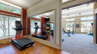 This well-equipped fitness center in Mansfield, TX is organized into distinct workout zones across a single floor, offering a comprehensive exercise environment. The cardio area features warm hardwood flooring, bold red accent walls, and large windows that flood the space with natural light and outdoor views of surrounding greenery. Treadmills, stationary bikes, and elliptical machines are arranged alongside wall-mounted mirrors that visually expand the room, while a mounted flat-screen TV adds convenience. A dedicated strength training zone transitions to textured carpet tile flooring and a more neutral color palette. This area is outfitted with a cable machine, multiple weight benches, a full dumbbell rack, and a cable/functional trainer system. Large windows continue throughout this section, maintaining the bright, airy atmosphere. An additional open area near the entrance provides space for functional training, featuring a bench and wall-mounted TV. Glass-paneled doors connect this zone to an adjacent room, adding visual openness. Elevated clerestory windows in the strength area allow additional natural light from above. The overall design balances energetic, motivating aesthetics in the cardio zone with a practical, focused layout in the strength area, creating a functional and inviting fitness facility.