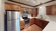 This single-floor space is thoughtfully designed with a cohesive aesthetic that balances warmth and functionality throughout. Rich wood trim and crown molding serve as unifying architectural details, present in both the open living area and kitchen.

The living area features dark hardwood-style flooring and neutral walls, creating a versatile backdrop. A visible thermostat and multiple outlets reflect practical planning throughout.

The kitchen is a standout feature, showcasing warm wood cabinetry with shaker-style doors and brushed hardware, complemented by light countertops and a full suite of stainless steel appliances, including a refrigerator, electric range, and built-in microwave. A double stainless steel sink with a matte black pull-down faucet adds a modern contrast, while pendant and track lighting illuminate the workspace effectively.

The bedroom offers plush carpeting, a window with an outdoor view of surrounding greenery, and a ceiling-mounted light fixture, providing a quiet and comfortable retreat.

The full bathroom is finished with tile flooring, a wood vanity, a large mirror, and a full tub-shower combination, maintaining the consistent warm-toned palette found throughout the space.