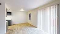 This two-story space is thoughtfully laid out across two distinct levels connected by a carpeted staircase. The main level features wood-look plank flooring throughout, lending a clean, cohesive aesthetic to the living and dining areas. A generous sliding glass door dressed with vertical blinds allows abundant natural light to fill the main living space, which also includes a separate entry door and a wall-mounted heater. Adjacent to the living area, a galley-style kitchen is outfitted with dark gray cabinetry, light speckled countertops, a black electric range, a refrigerator, and a stainless steel sink beneath a skylight-style ceiling fixture, offering ample storage and counter space. A full bathroom on the lower level includes wood-look flooring, a tub-shower combination, a dark-cabinet vanity with a white sink, and a mirrored medicine cabinet with bar lighting above. The upper level transitions to plush carpet throughout the bedrooms, each offering white walls, natural window light, and ceiling fans for comfort. One carpeted upper room features dual doorways leading to additional spaces. The overall palette of crisp white walls, neutral flooring, and dark fixture accents creates a clean, functional environment throughout.
