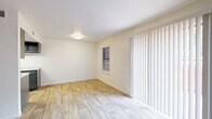 This two-story space is thoughtfully laid out across two distinct levels connected by a carpeted staircase. The main level features wood-look plank flooring throughout, lending a clean, cohesive aesthetic to the living and dining areas. A generous sliding glass door dressed with vertical blinds allows abundant natural light to fill the main living space, which also includes a separate entry door and a wall-mounted heater. Adjacent to the living area, a galley-style kitchen is outfitted with dark gray cabinetry, light speckled countertops, a black electric range, a refrigerator, and a stainless steel sink beneath a skylight-style ceiling fixture, offering ample storage and counter space. A full bathroom on the lower level includes wood-look flooring, a tub-shower combination, a dark-cabinet vanity with a white sink, and a mirrored medicine cabinet with bar lighting above. The upper level transitions to plush carpet throughout the bedrooms, each offering white walls, natural window light, and ceiling fans for comfort. One carpeted upper room features dual doorways leading to additional spaces. The overall palette of crisp white walls, neutral flooring, and dark fixture accents creates a clean, functional environment throughout.