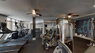This single-floor fitness center at 1300 Tribute Center Dr in Raleigh, NC offers a comprehensive workout facility designed to accommodate a wide range of exercise needs. The space is organized into several distinct zones, each purpose-built for specific types of training. The cardio area features a row of Matrix treadmills and a bank of elliptical machines, all positioned along a wall of floor-to-ceiling windows that allow abundant natural light and views of the surrounding outdoor landscape. A dedicated strength training zone houses a robust selection of Hoist multi-station cable machines and selectorized equipment, providing options for full-body resistance workouts. A separate free weights area contains multiple racks of dumbbells in graduated sizes, complemented by a wall-mounted flat-screen television for entertainment. An additional zone accommodates upright and recumbent stationary bikes, also monitored by wall-mounted screens. Throughout the facility, rubber sport flooring provides traction and impact absorption, while ceiling fans and recessed lighting ensure a comfortable environment. Decorative columns with crown molding detail lend a polished, refined aesthetic to the otherwise functional space, maintaining a balance between practicality and design quality.